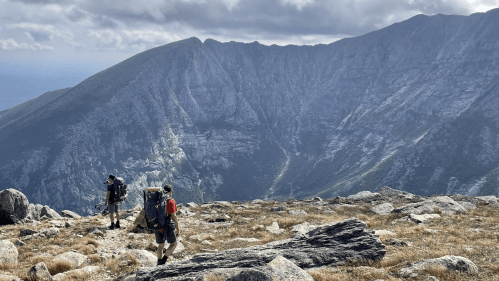Two hikers with backpacks trek along a rocky trail, surrounded by mountains and a cloudy sky.