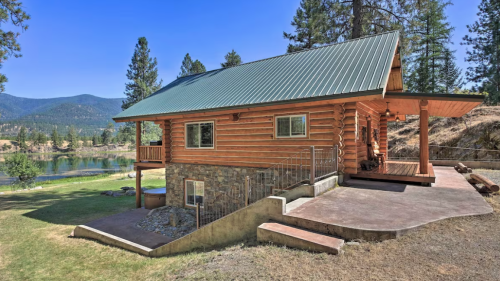 A rustic log cabin with a green metal roof, surrounded by trees and a scenic view of a lake and mountains.