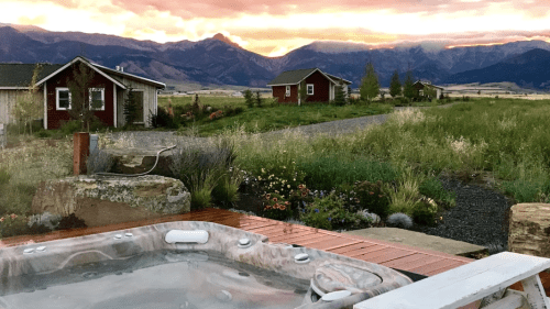 A hot tub on a wooden deck with a scenic view of mountains and cabins at sunset, surrounded by lush greenery.