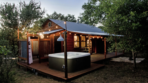 Cozy cabin with string lights and a hot tub on a wooden deck, surrounded by trees in a serene outdoor setting.