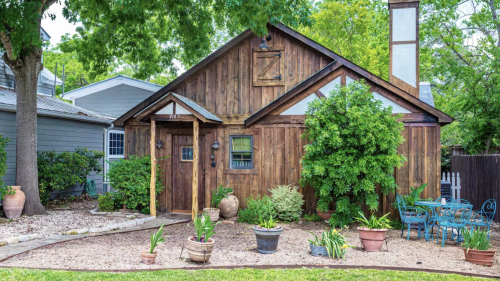A charming wooden house surrounded by greenery and potted plants, with a small patio and outdoor seating area.