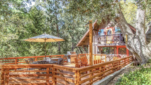 A cozy cabin with a wooden deck, outdoor seating, and an umbrella, surrounded by lush trees.