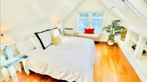 Bright, cozy bedroom with a large bed, wooden floors, and a view of the water through a window.