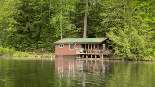 A rustic cabin by a calm lake, surrounded by lush green trees and reflecting in the water.