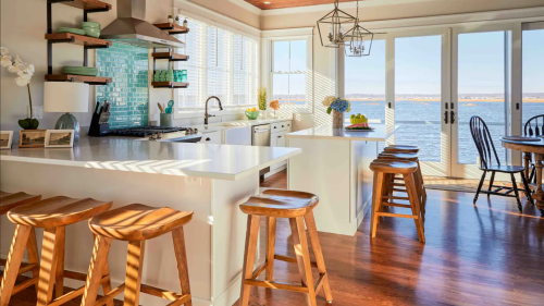 Bright kitchen with white cabinets, wooden stools, and a view of the water through large windows. Sunlight casts shadows.