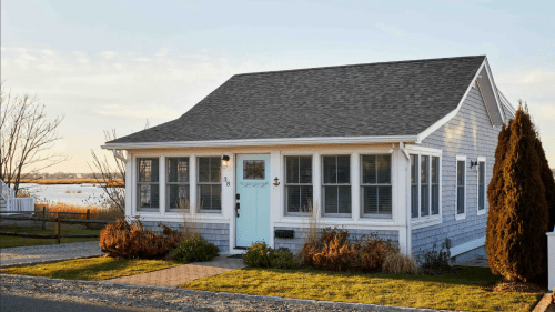 A charming blue cottage with a light blue door, surrounded by greenery and a view of water in the background.