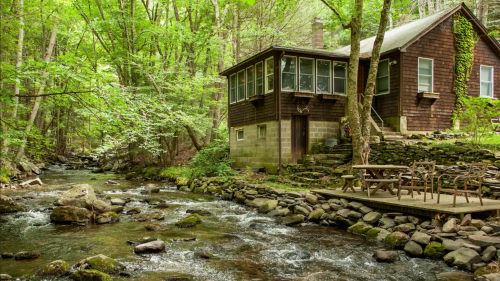 A rustic cabin by a flowing stream, surrounded by lush green trees and rocky terrain.