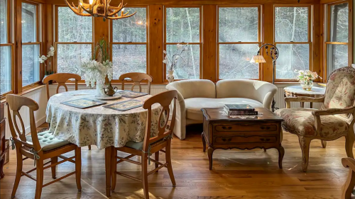 Cozy sunroom with wooden accents, a floral tablecloth, a white sofa, and large windows overlooking nature.