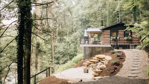 A cozy log cabin nestled in a forest, surrounded by trees and featuring a stone patio with a fire pit.
