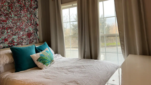 Cozy bedroom with a colorful abstract wall art, a bed with decorative pillows, and large windows with curtains.