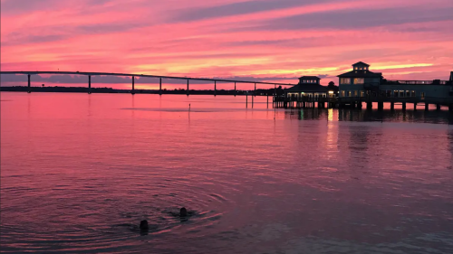 A vibrant sunset casts pink and purple hues over calm water, with two swimmers and a pier in the background.