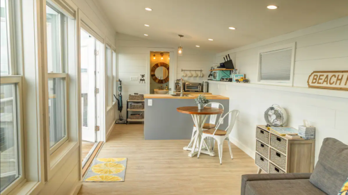 Bright, modern interior of a beach house with a dining area, kitchen, and cozy seating. Large windows let in natural light.