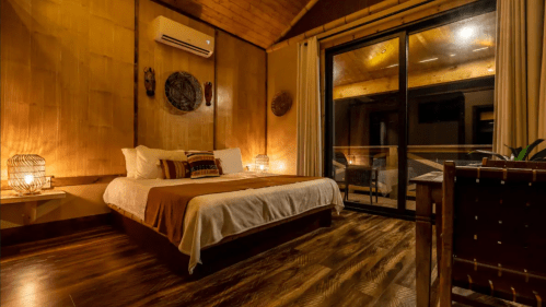 Cozy bedroom with wooden walls, a large bed, and glass doors leading to a balcony, softly lit for a warm ambiance.