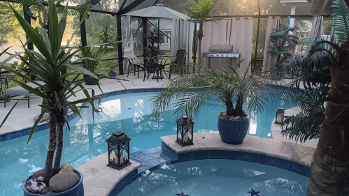 A serene backyard pool area with palm trees, lanterns, and a grill, surrounded by a cozy seating arrangement.