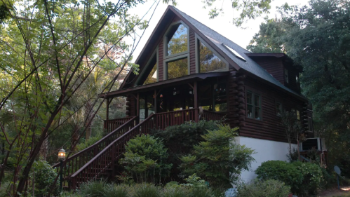 A charming log cabin with large windows, surrounded by trees and greenery, featuring a welcoming front porch.