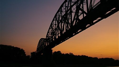 Silhouette of a bridge against a colorful sunset over a river, with trees lining the shore.