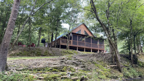 A cozy cabin nestled among trees by a stream, featuring a porch and outdoor seating.