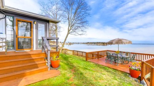 A lakeside view featuring a wooden deck, patio furniture, and a tree, with calm water and blue skies in the background.