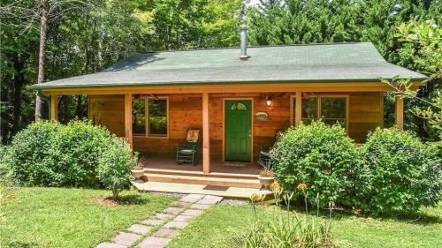 A cozy wooden cabin with a green door, surrounded by lush greenery and a stone pathway.