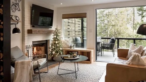 Cozy living room with a fireplace, Christmas tree, and large windows overlooking a forested area. Modern decor and furniture.