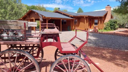 A rustic log cabin with a gray roof, surrounded by trees and a gravel path, featuring a vintage red wagon in the foreground.
