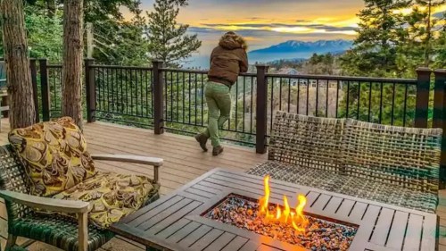 A person leans on a railing, overlooking a scenic sunset with mountains, surrounded by trees and a cozy outdoor seating area.