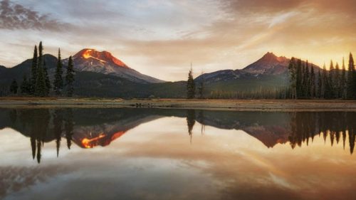 Sunset over two mountains reflected in a calm lake, surrounded by evergreen trees and a colorful sky.