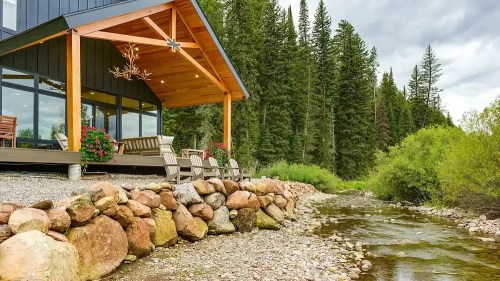 A modern cabin by a rocky riverbank, surrounded by lush trees and greenery under a cloudy sky.