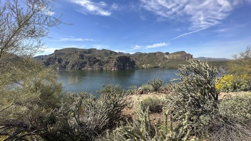 Scenic view of a calm lake surrounded by rocky hills and desert vegetation under a blue sky with wispy clouds.