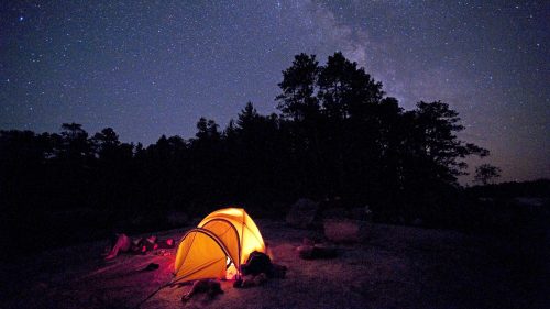 A glowing yellow tent under a starry night sky, surrounded by trees and a peaceful campsite.