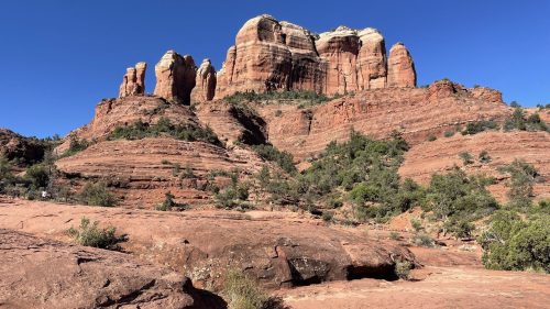 Red rock formations rise against a clear blue sky, surrounded by green vegetation in a desert landscape.