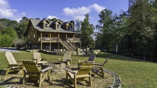 A cozy log cabin surrounded by trees, featuring a fire pit with wooden chairs in the foreground.