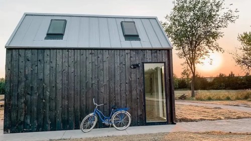 A modern black wooden house with a metal roof, a bicycle parked outside, and a sunset in the background.
