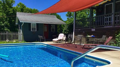 A sunny backyard with a blue pool, lounge chairs, and a shaded area under a red canopy. Green trees in the background.