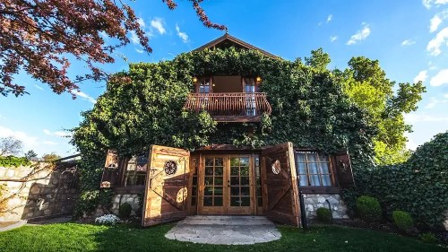 A charming wooden house covered in greenery, featuring open double doors and a balcony under a blue sky.