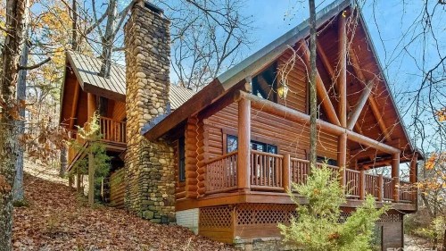 A rustic log cabin with a stone chimney, surrounded by autumn foliage and trees.