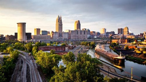 Cleveland skyline at sunset, featuring buildings, a river, and a cargo ship, surrounded by greenery and rail tracks.