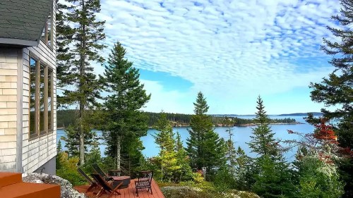 A scenic view of a lake surrounded by trees, with a house and deck in the foreground under a cloudy sky.