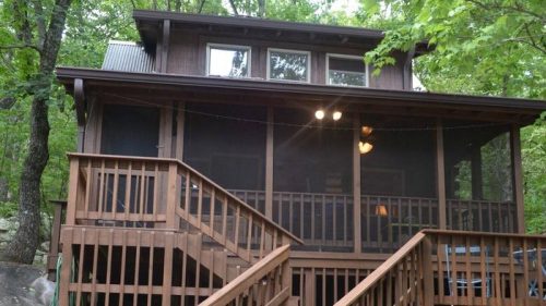 A wooden cabin with a screened porch, surrounded by trees and rocks, featuring stairs leading to the entrance.