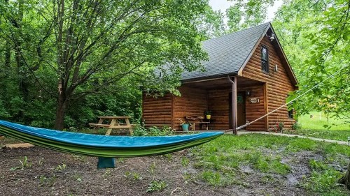 A cozy log cabin surrounded by trees, with a hammock and picnic table in the foreground.