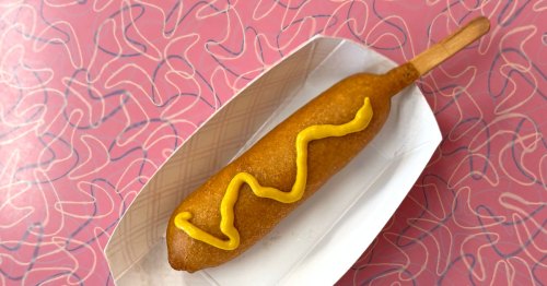 A corn dog with yellow mustard sits in a dish on a pink table.