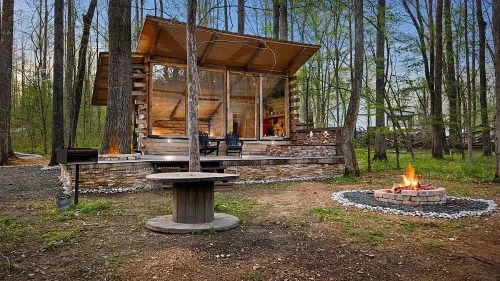 A cozy cabin in the woods with a stone fire pit and seating area, surrounded by trees and a serene landscape.