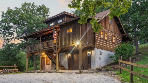 A rustic wooden house with a balcony, surrounded by trees and a gravel driveway, featuring an American flag.