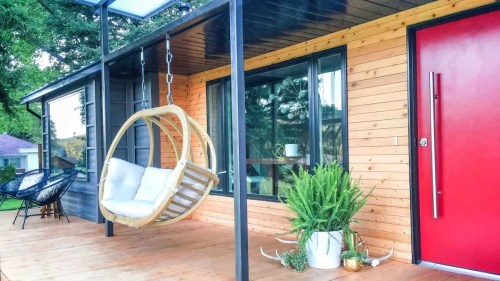 A modern porch with a hanging swing chair, wooden deck, large windows, and a vibrant red door. Green plants add a cozy touch.