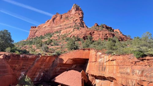 A striking red rock formation rises against a clear blue sky, surrounded by greenery and rugged terrain.