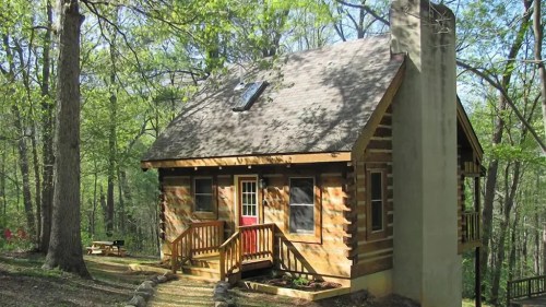 A cozy log cabin surrounded by trees, featuring a stone chimney and a welcoming front porch.