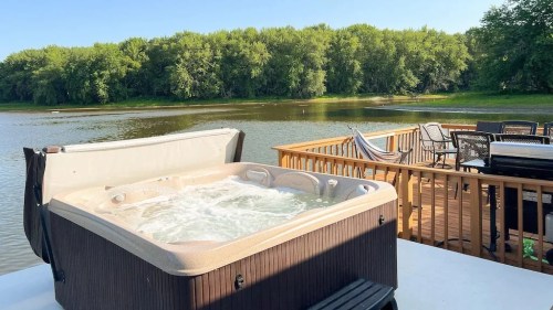 A hot tub on a deck overlooking a calm river surrounded by lush green trees under a clear blue sky.
