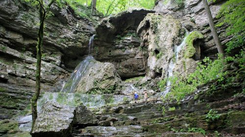 A serene waterfall cascades down rocky cliffs, surrounded by lush greenery and two people exploring the area.