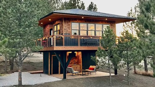 A modern two-story cabin surrounded by pine trees, featuring a balcony and outdoor seating area.