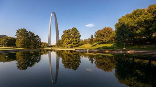 The Gateway Arch reflects in a calm pond, surrounded by green trees and a clear blue sky.